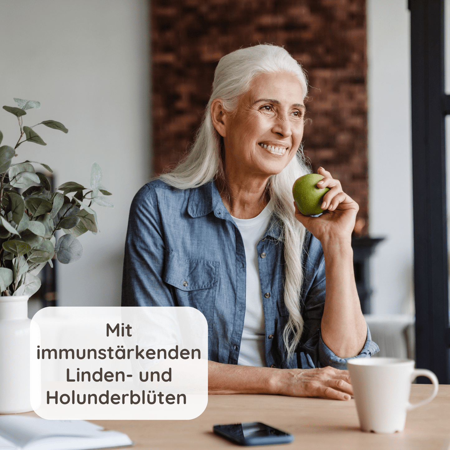 Ältere Frau sitzt lächelnd am Tisch und hält einen grünen Apfel mit folgendem Text: Mit immunstärkenden Linden- und Holunderblüten.