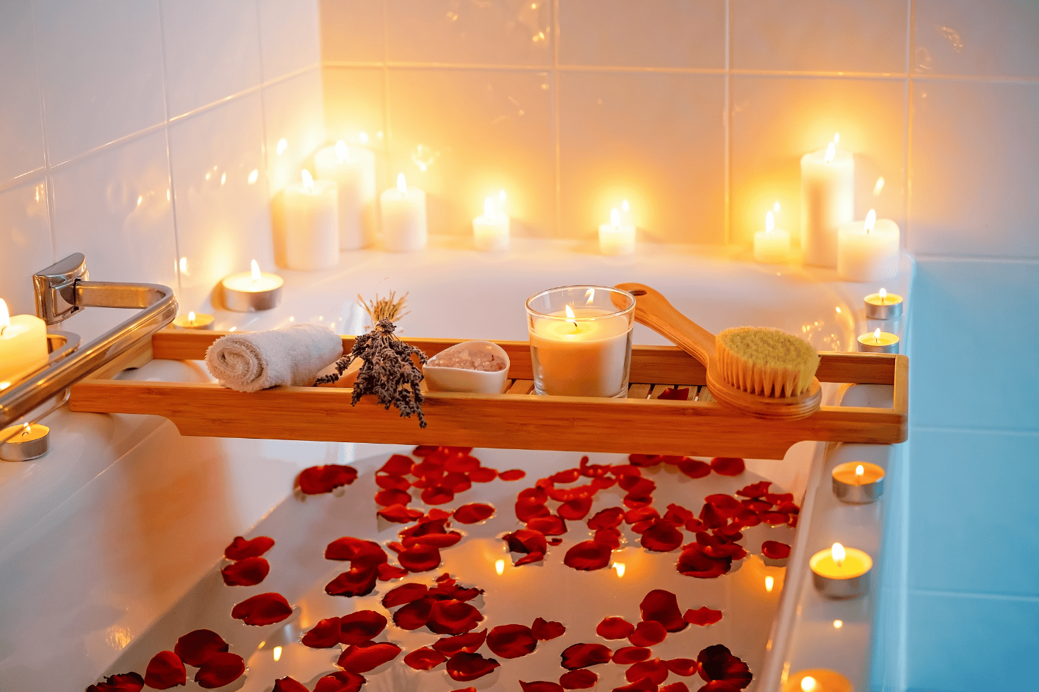 Badewanne mit eingelassenem Wasser, umgeben von brennenden Kerzen. Auf dem Badewasser schwimmen einige Rosenblätter.