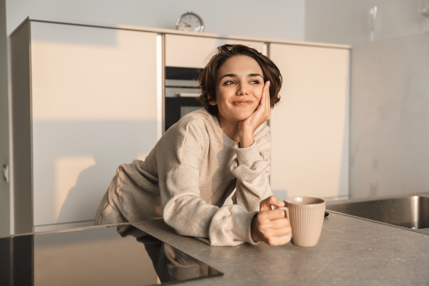 Foto von einer jungen Frau mit Tasse Tee in der Hand, die sich über ihre Kücheninsel beugt und fröhlich aussieht.