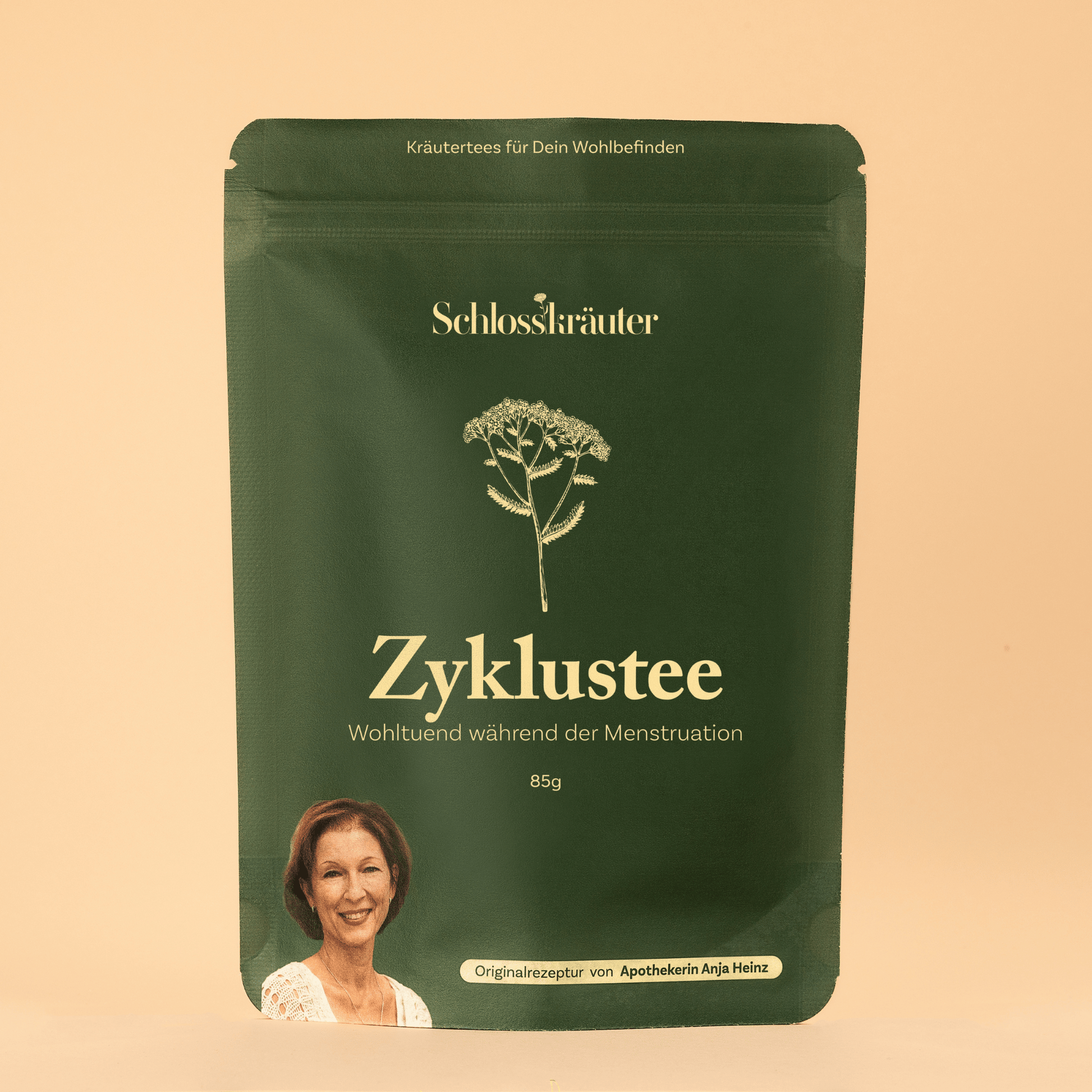 Grüne Teeverpackung mit der Aufschrift „Zyklustee – Wohltuend während der Menstruation“, vor beigem Hintergrund, mit Illustration einer Heilpflanze und einem Portraitfoto von Apothekerin Anja Heinz.