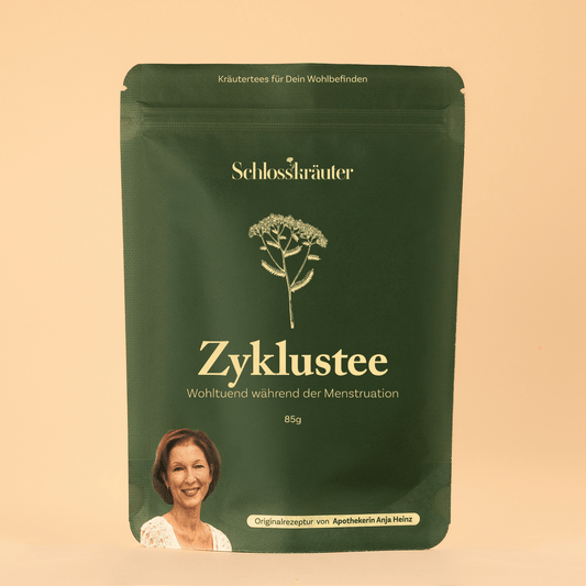 Grüne Teeverpackung mit der Aufschrift „Zyklustee – Wohltuend während der Menstruation“, vor beigem Hintergrund, mit Illustration einer Heilpflanze und einem Portraitfoto von Apothekerin Anja Heinz.