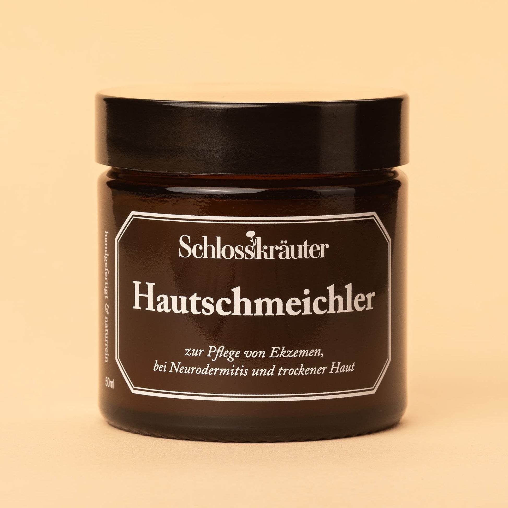 Braunes Glas mit schwarzem Deckel und Etikett „Hautschmeichler – zur Pflege bei Neurodermitis, Ekzemen und trockener Haut“, vor beigem Hintergrund.
