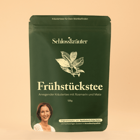 Grüne Teeverpackung mit der Aufschrift „Frühstückstee - Anregender Kräutertee mit Rosmarin und Mate“, vor beigem Hintergrund