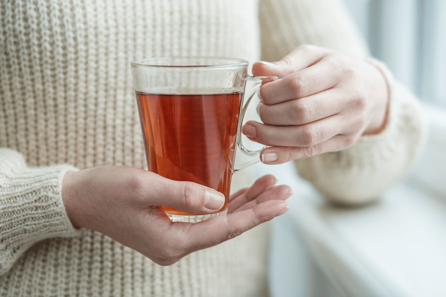 Eine Person hält eine durchsichtige Tasse mit Kräutertee. Sie trägt einen beigen Strickpullover. Man sieht die Tasse und die Hände der Person, die sie halten.