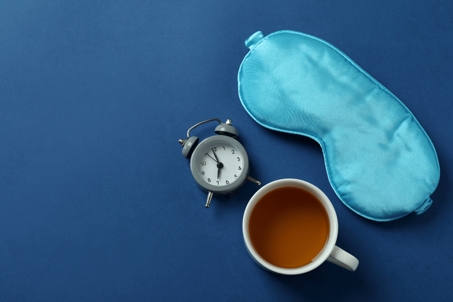 Tasse Tee, Wecker und eine hellblaue Schlafmaske liegen auf dunkelblauem Hintergrund, arrangiert als Stillleben.